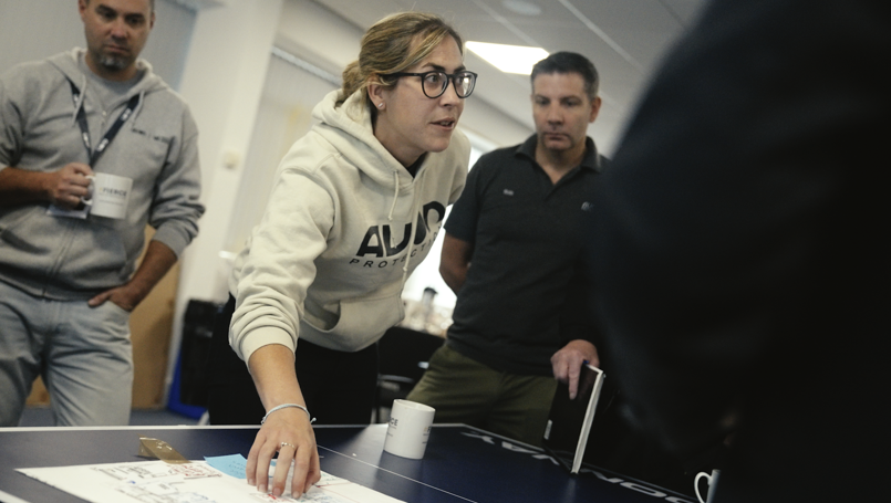 A female employee in an "Avon Protection" hoodie leans over a table to explain a project plan to two male colleagues during a team meeting.