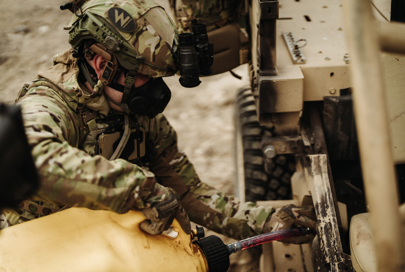 A person in multicam tactical gear and a respirator mask refueling a tan military vehicle using a yellow fuel canister.