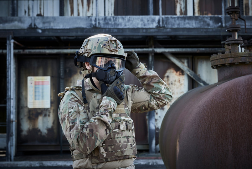 A tactical operator in multicam gear adjusting a full-face respirator mask while standing next to a large, rusted industrial tank.