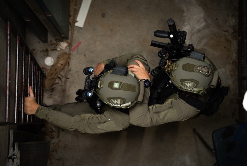 An overhead shot of two tactical operators in green uniforms and ballistic helmets stack positioned against a metal door in a dimly lit environment.