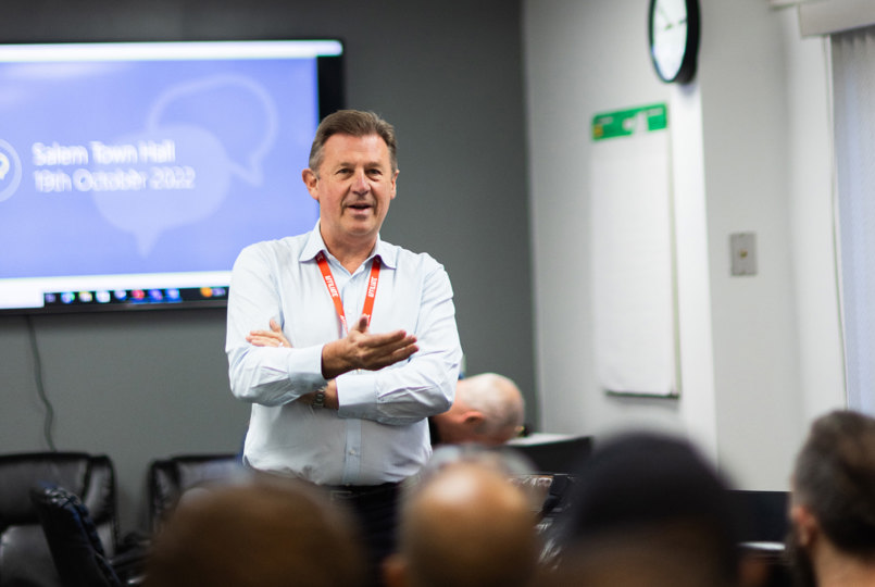 A man in a light blue shirt and red lanyard gestures while speaking at a "Salem Town Hall" meeting in a conference room.