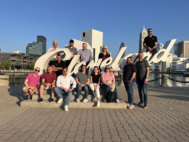 A large group of colleagues posing for a photo around the white script "Cleveland" sign at Voinovich Bicentennial Park, with the city skyline and harbor in the background.