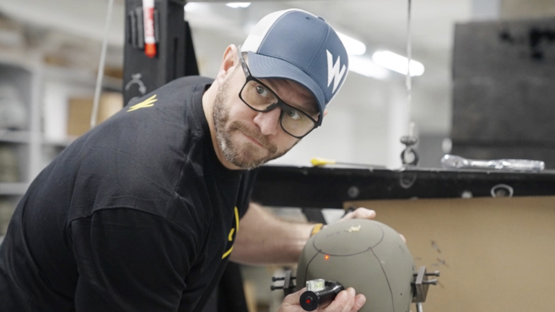 Technician using a laser alignment tool on a tactical ballistics helmet during safety testing in a workshop.