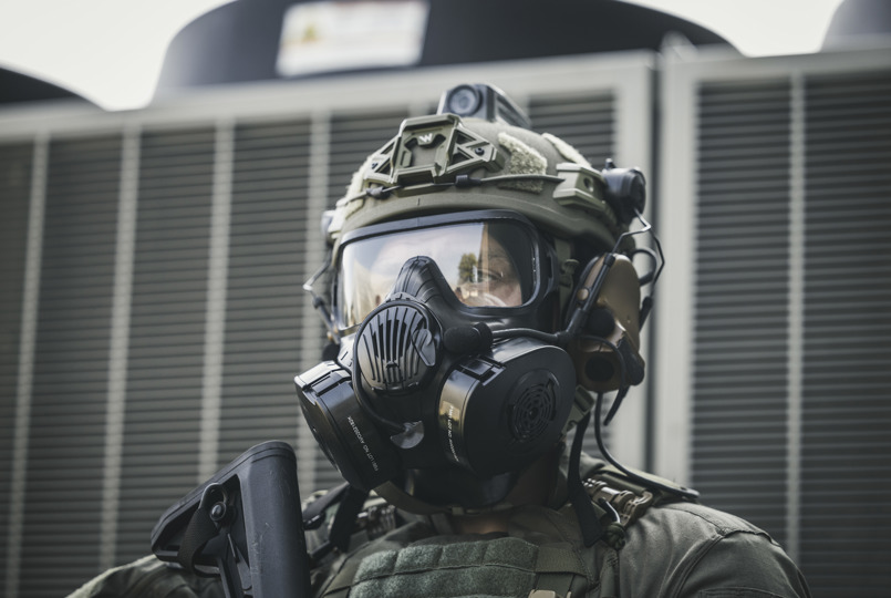 A close-up of a person in tactical gear wearing a full-face respirator mask, a ballistic helmet with a mounted camera, and communication headsets.