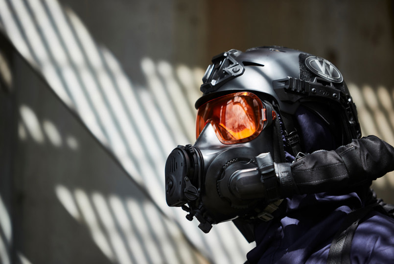 A close-up profile of an operator wearing a matte black tactical helmet and a respirator mask with high-visibility orange tinted lenses.