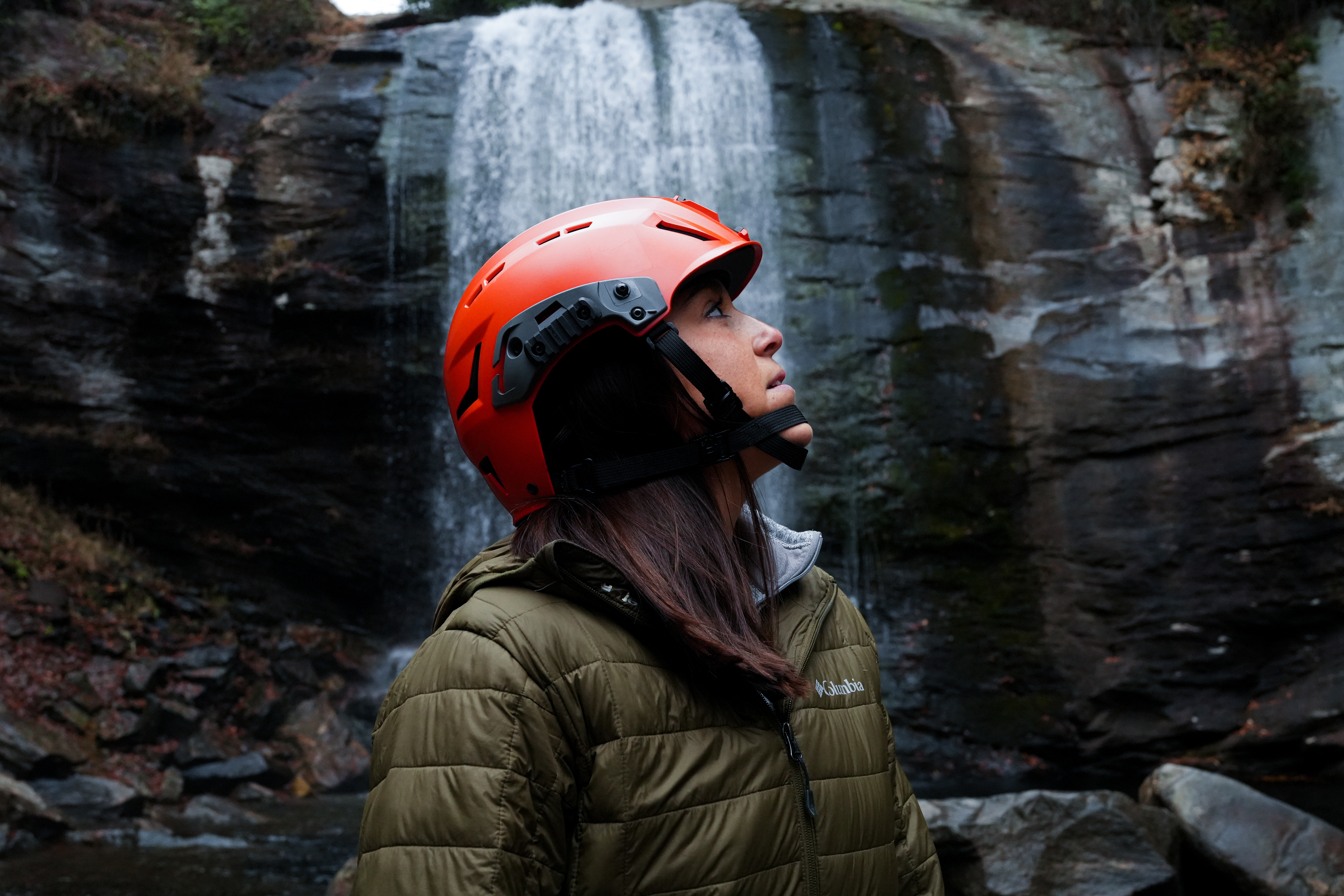 A woman in an olive green puffer jacket and a bright orange safety helmet looking up at a waterfall cascading over dark rocks.