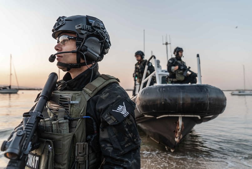 A tactical officer in a helmet and headset standing on a shore near a rigid inflatable boat (RIB) with other team members.