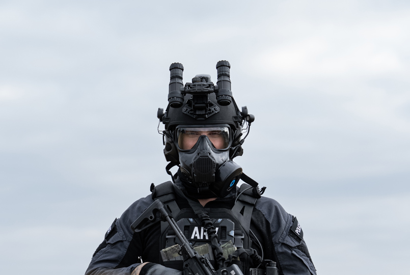 A tactical officer in a black uniform and respirator mask holding a rifle while standing in a field of tall grass.