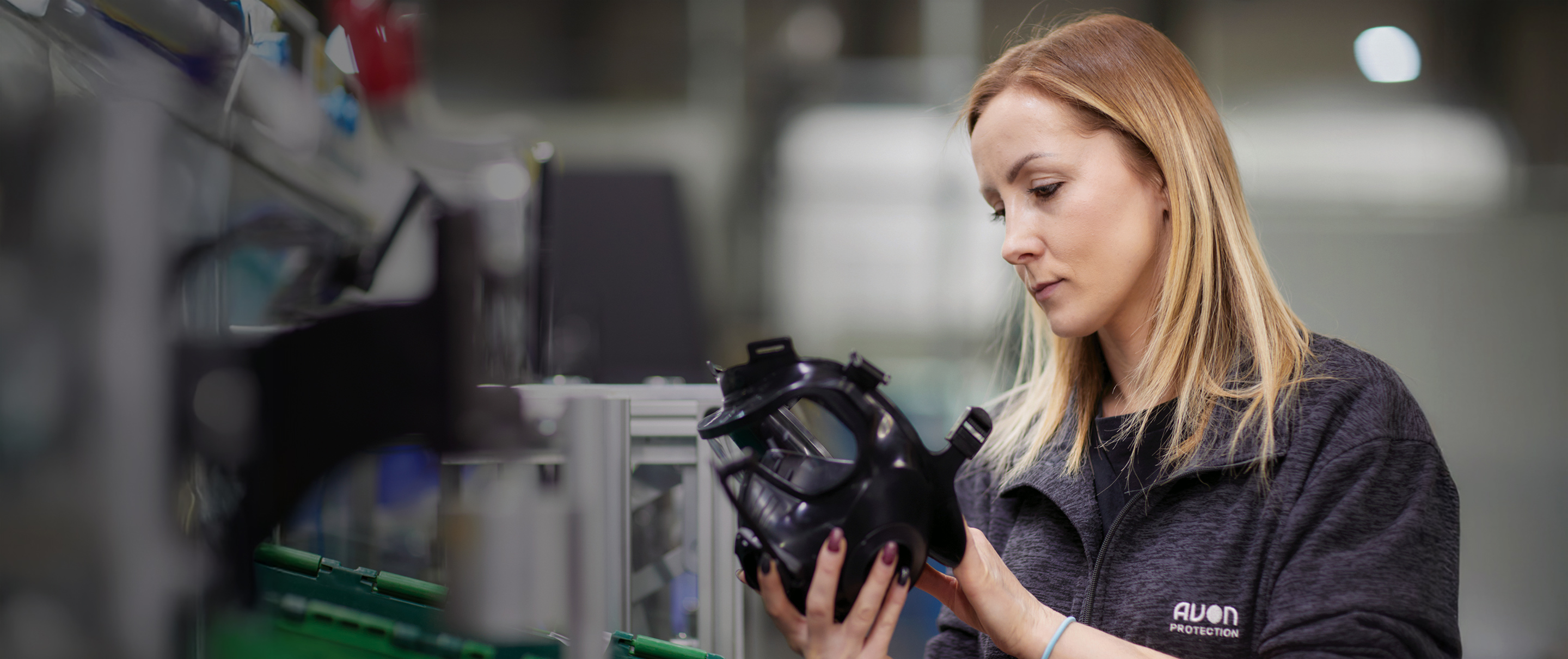 A female worker in an "Avon Protection" branded fleece inspecting a black tactical respirator mask in a factory setting.