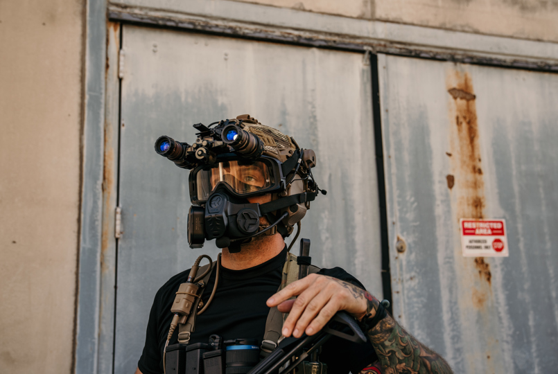 A tactical operator wearing a respirator mask and a ballistic helmet with dual-tube night vision goggles (NVGs) standing in front of a weathered industrial door.