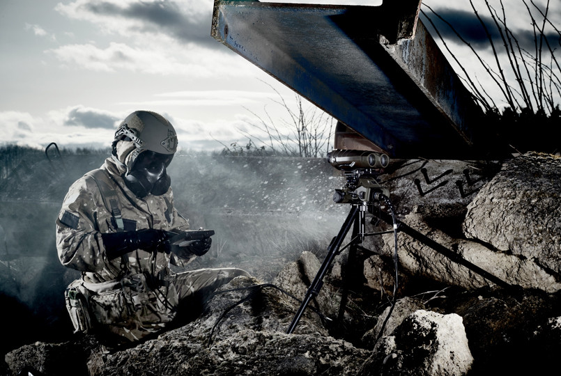 A person in military camouflage and a gas mask operating a handheld device next to a tripod-mounted camera in a rocky landscape.
