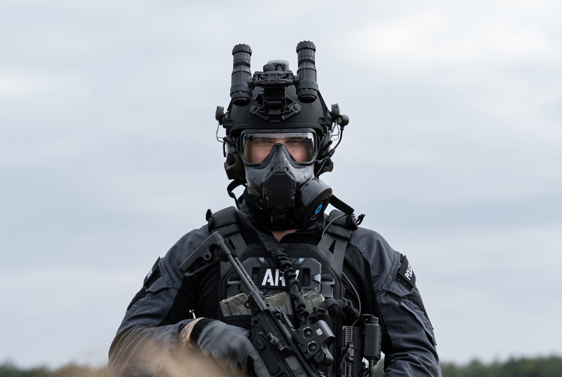 A tactical operator in a grey uniform holding a rifle and wearing a full-face respirator mask and a ballistic helmet with stowed night vision goggles.