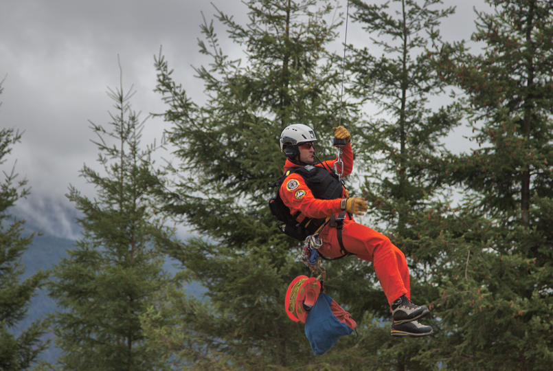 A search and rescue technician in a bright orange jumpsuit and white helmet rappels from a cable against a backdrop of evergreen trees.