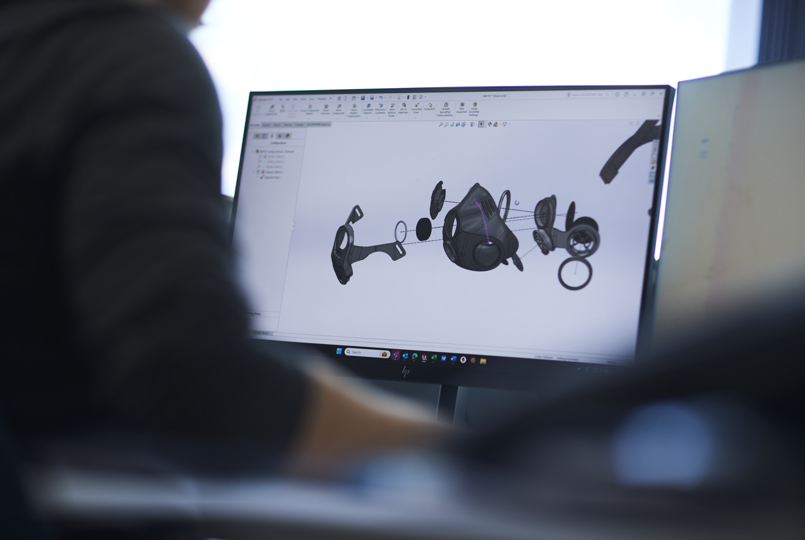A computer monitor displaying a 3D CAD (Computer-Aided Design) model of a disassembled respirator mask being designed in an office.