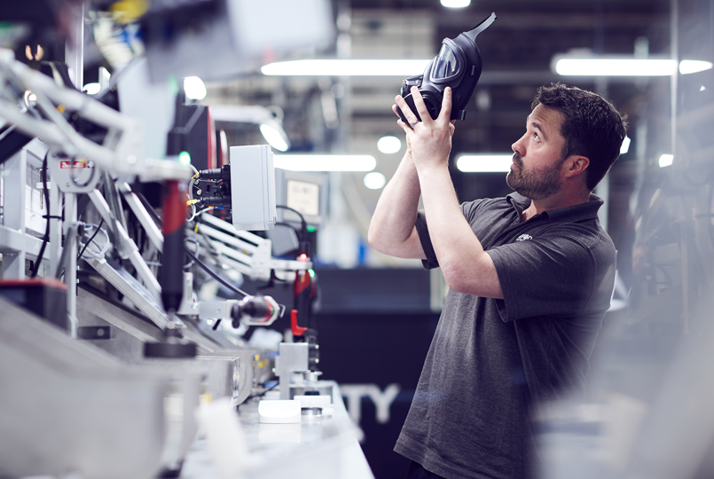 A male worker in a grey polo shirt inspecting a black respirator mask in a brightly lit industrial manufacturing facility.