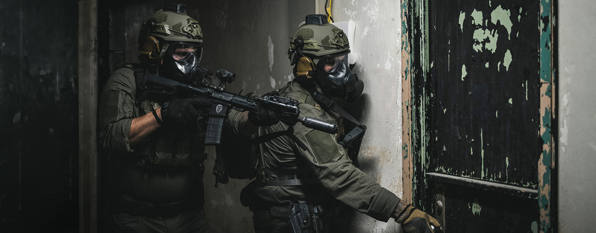 Two soldiers wearing helmets and respirators leaning on wall with firearms