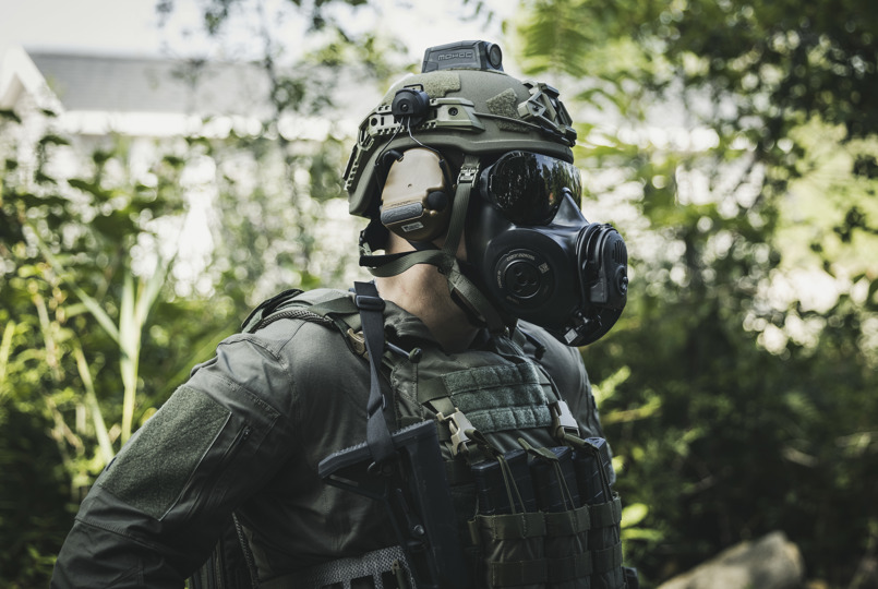 Side profile of a tactical operator wearing an olive green helmet, a black respirator mask, and a tactical vest in a wooded area.