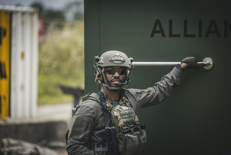 A tactical operator in an olive green uniform and tan ballistic helmet standing next to a large green structure labeled "ALLIANCE."