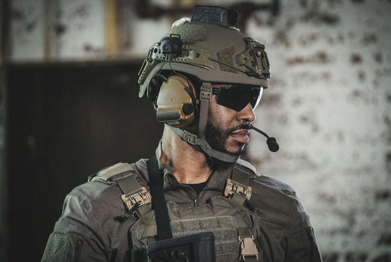 A close-up profile of a man in tactical gear, wearing a tan ballistic helmet with integrated communication headsets, a boom microphone, and dark safety glasses.